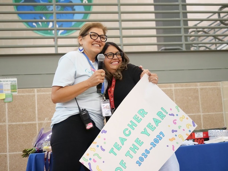 Two women hugging each other, the woman is hold a microphone while the other woman is carrying a sign that says "Teacher of the Year! 2025-2026."