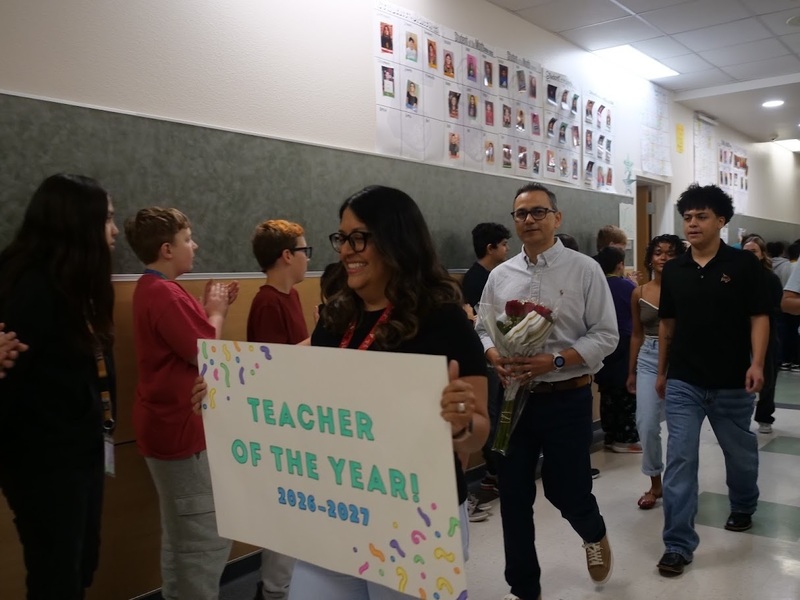 A group of people in a classroom hallway with a woman carrying a sign that says "Teacher of the Year! 2025-2026"  with people following behind.