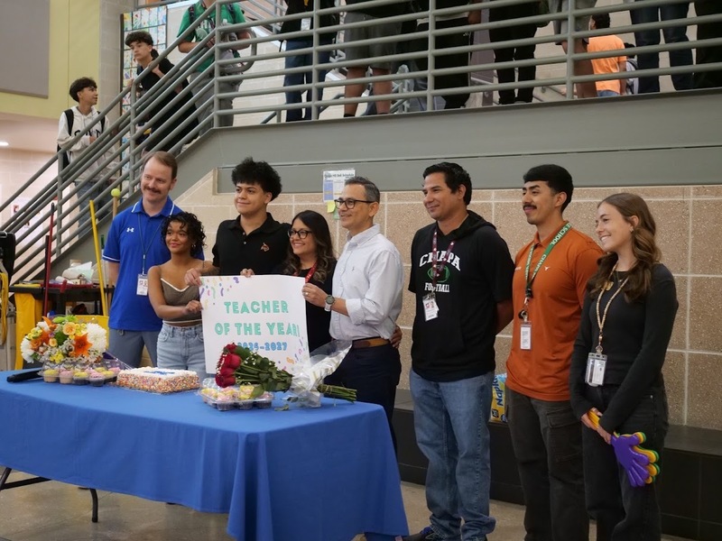 A group of people standing a behind a table while posing together for a group photo with the teacher of the year carrying a sign that says "Teacher of the Year! 2025-2026."