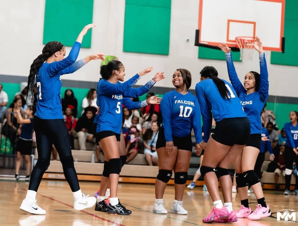 High school girls' volleyball team members wearing blue and black "Falcons" jerseys high-fiving and celebrating on the court during a game.