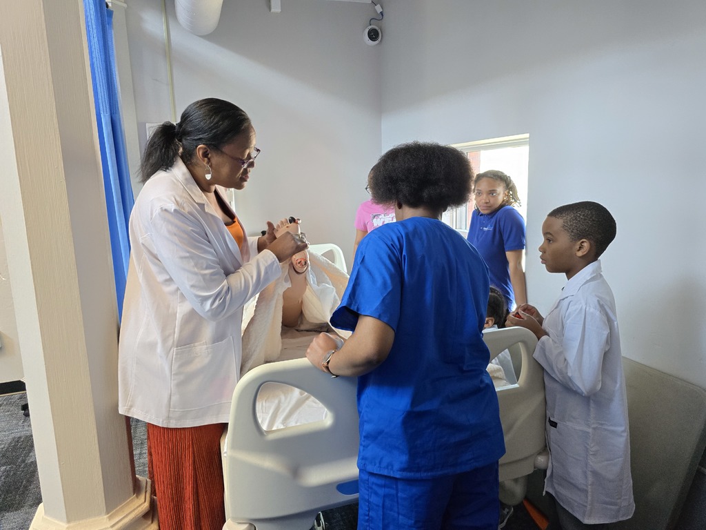 Students gathered around a hospital bed  learning from a teacher demonstrating with a manikin