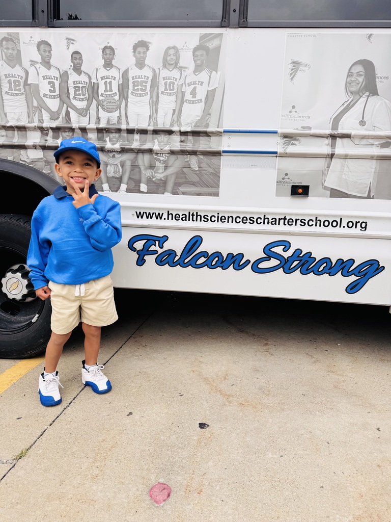 Young boy named Daniel wearing a blue polo, blue cap, and shorts, smiling and making a peace sign in front of a Health Sciences Charter School bus with the words "Falcon Strong" and a picture of a graduate on it.