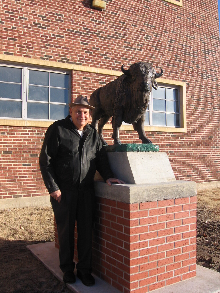 Man in black jacket standing next to bison statue on brick base.