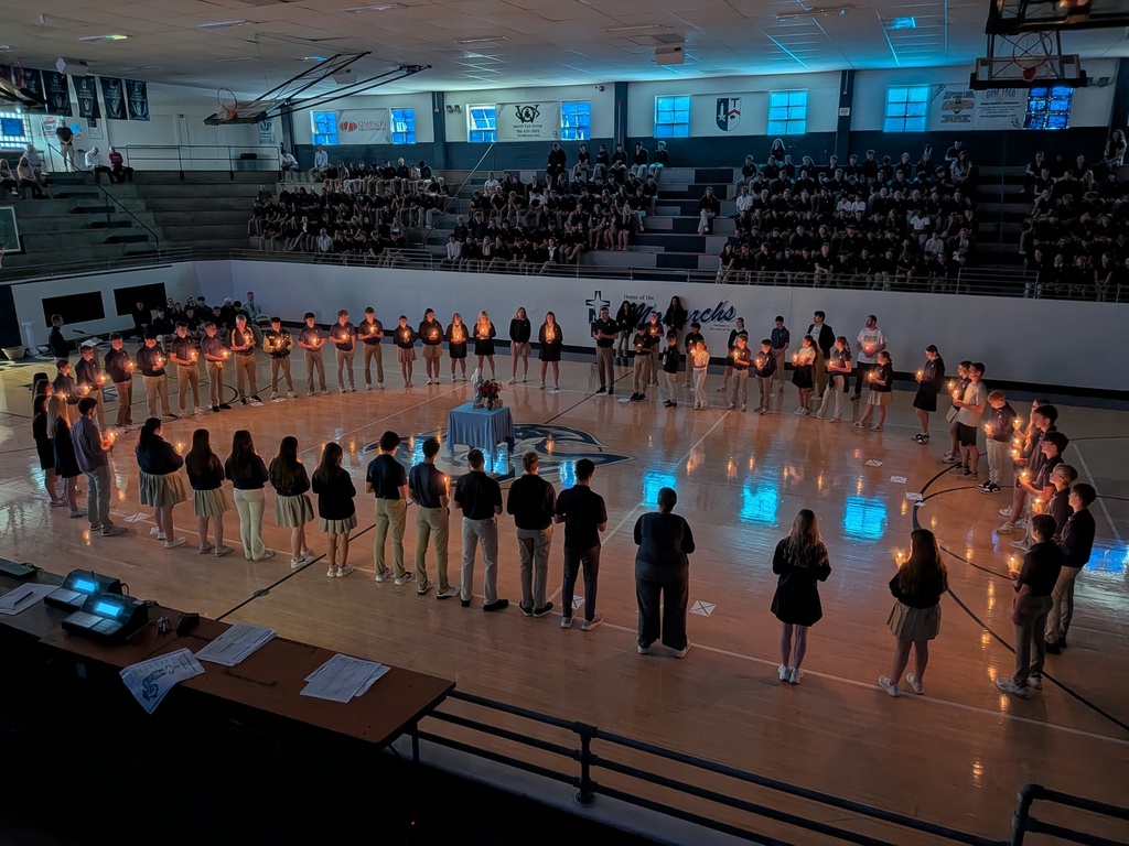 Students and teachers sit in the bleachers and stand on the floor holding candles