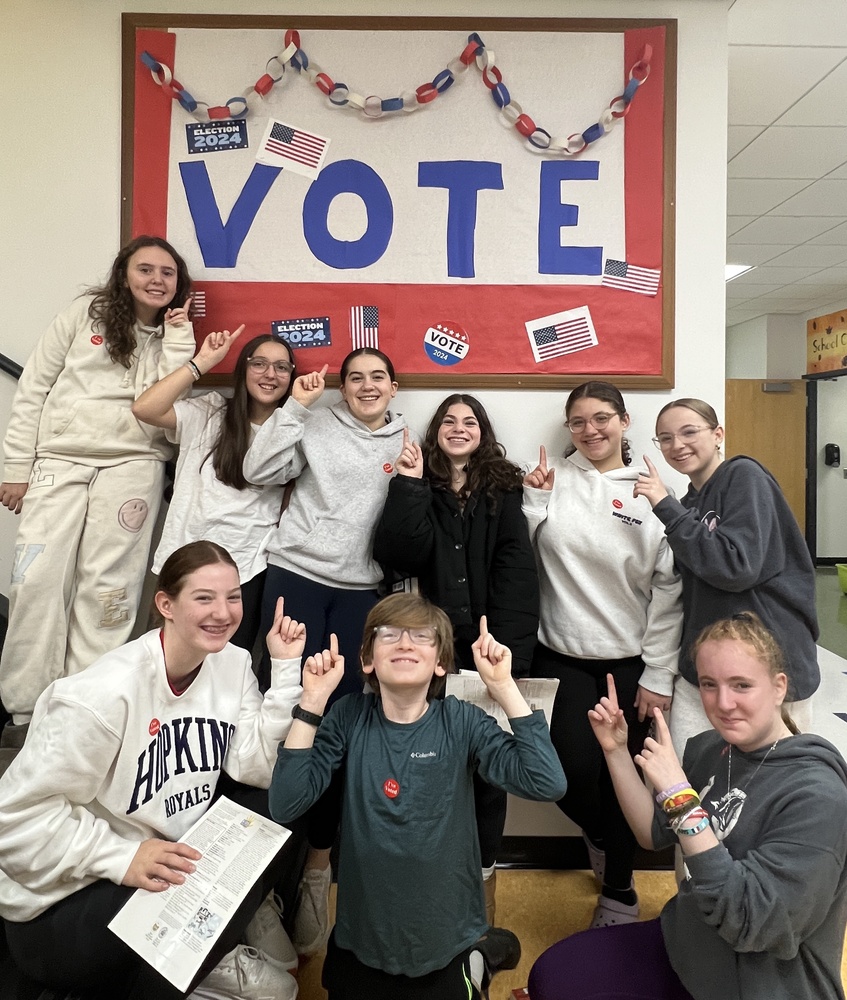 eighth graders wear I Voted stickers and pose pointing at bulletin board decorated for election day that says vote and has american flags and red white and blue