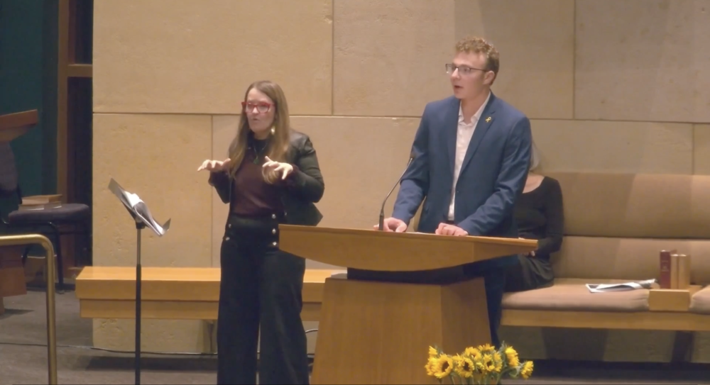 Alum Micah speaks at the pulpit on the Adath bimah. ASL interpreter interprets.