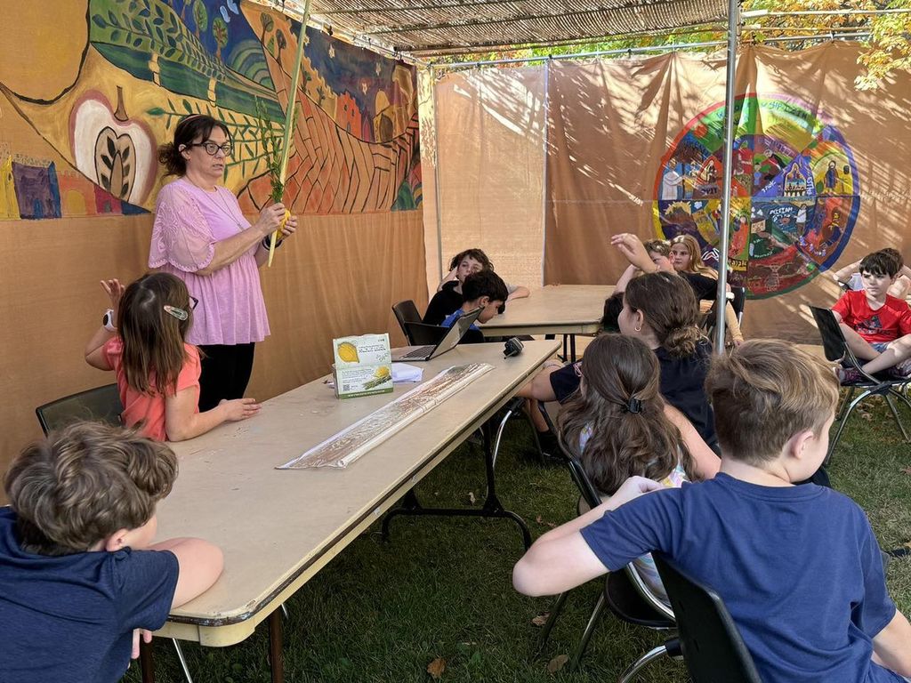 HaMorah Esther holds lulav and etrog to teach students in sukkah. two students are raising their hands