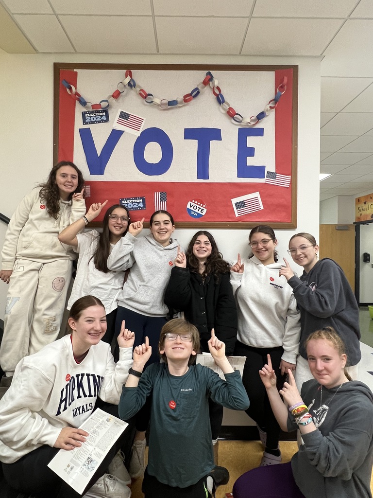 eighth graders wear I Voted stickers and pose pointing at bulletin board decorated for election day that says vote and has american flags and red white and blue