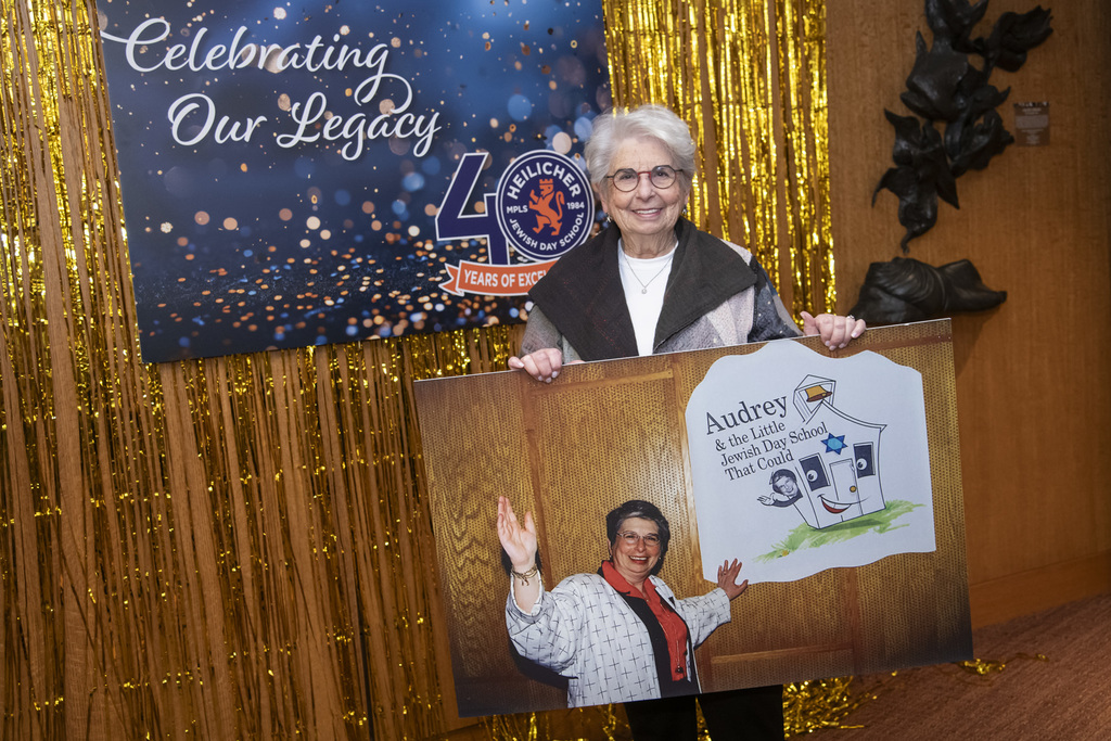 Honoree Audrey Goldfarb poses with photo of her from her time at Heilicher with a sign that says Audrey and the little jewish day school that could