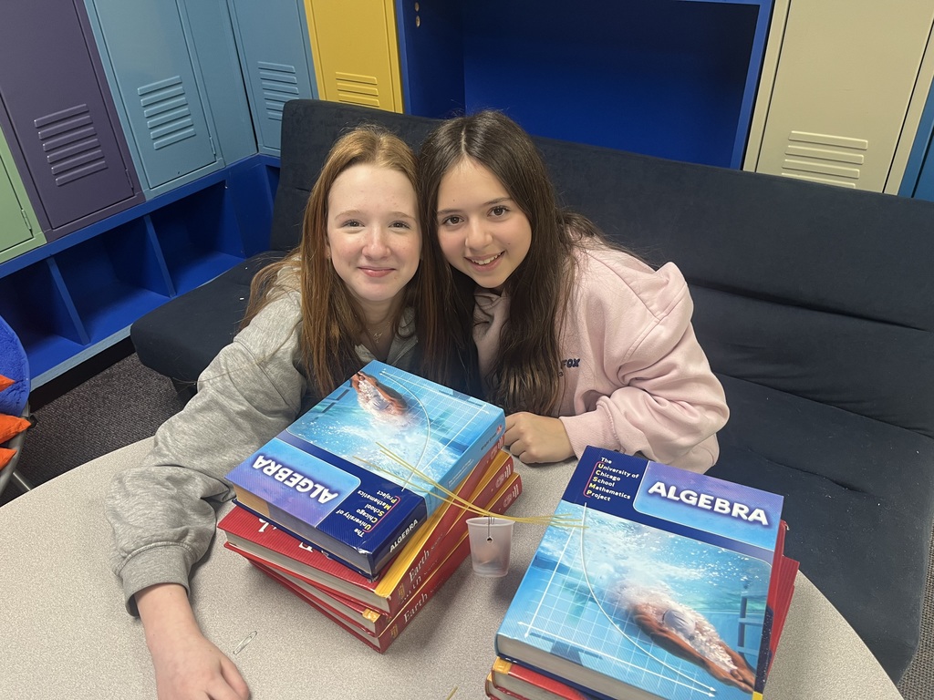 two seventh-grade girls with their spaghetti bridge held up by textbooks supports bucket of pennies
