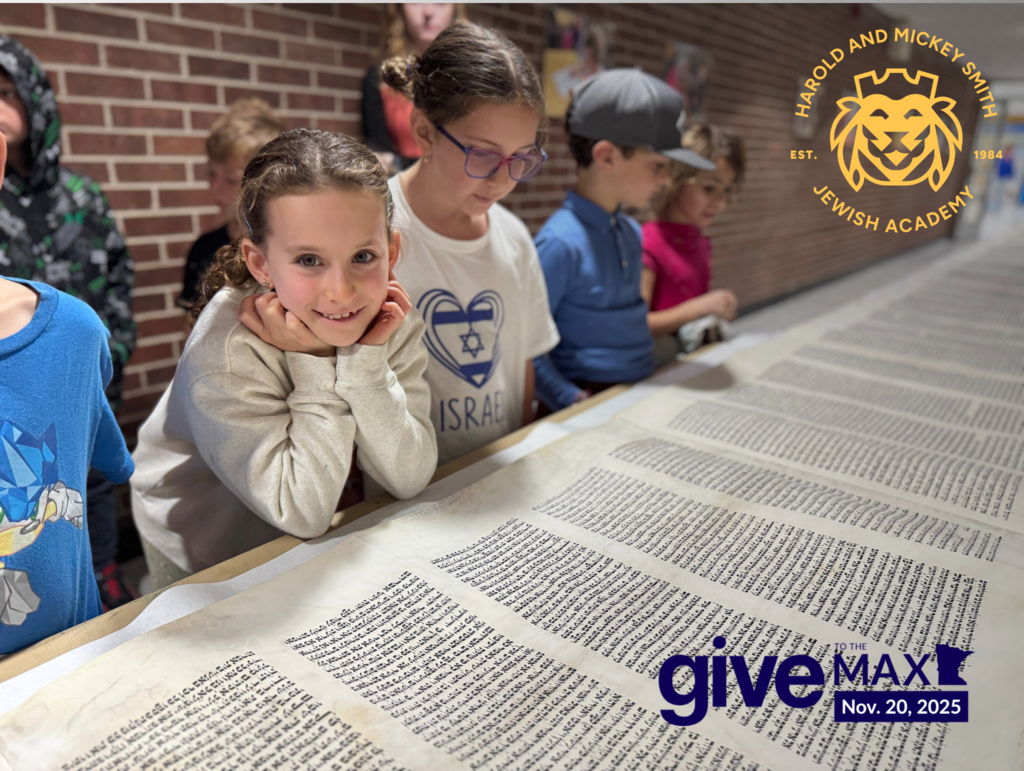 one student smiles at camera as class looks over rolled out Torah. Give to the Max Nov. 20, 2025 Harold and Mickey Smith Jewish Academy