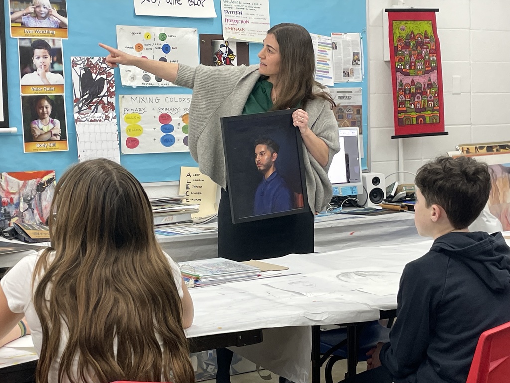 visiting artist/SJA parent points out of frame and holds portrait: man from the chest up, sitting in wooden chair wearing blue collared shirt. Black hair and stubble.
