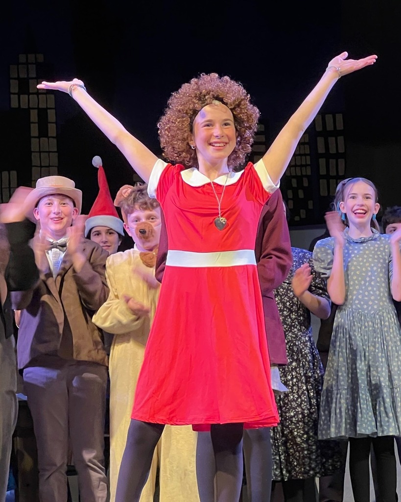 Annie poses with arms up and out as the rest of the cast claps