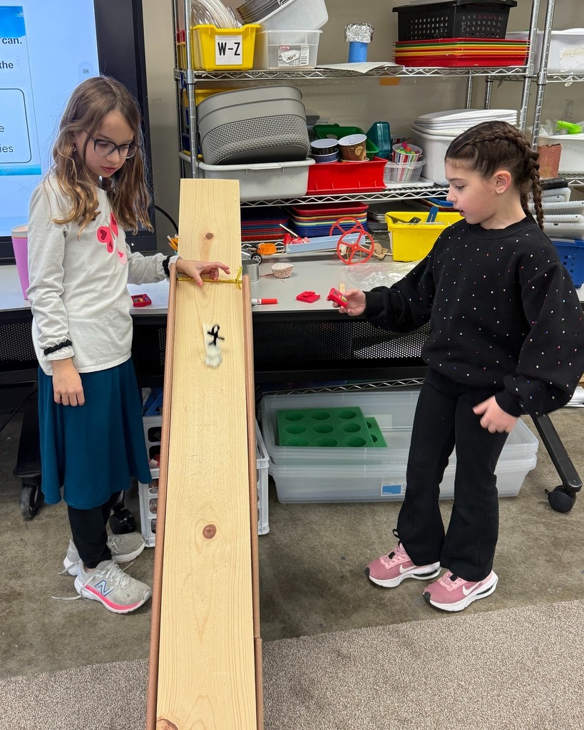 student releases snowboard creation down wooden slope while another student holds timer