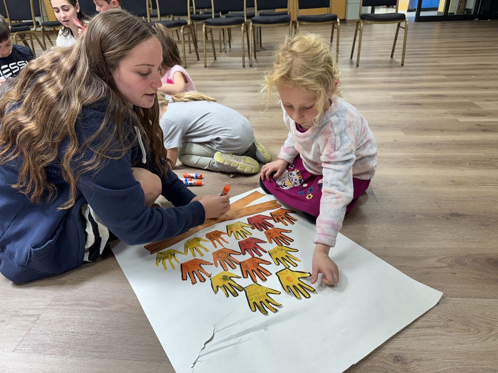 bat sherut helps student glue yellow, orange, red paper hands to poster to create bonfire image
