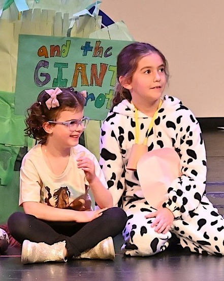 students sit onstage in costume, one as a horse and one as a cow