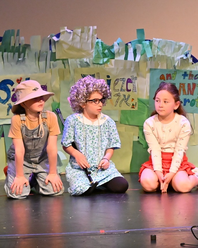 kids sit on stage, one as an elder farmer man, one as an elder woman, and one as a girl