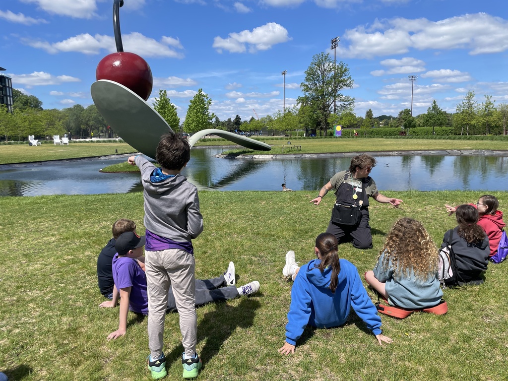 fifth graders and tour guide sit at edge of pond with Spoonbridge and Cherry sculpture 