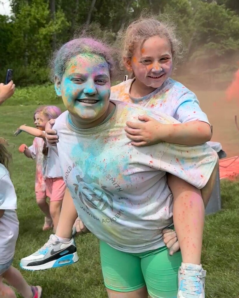 older student gives younger student piggyback ride; they are covered in colorful chalk
