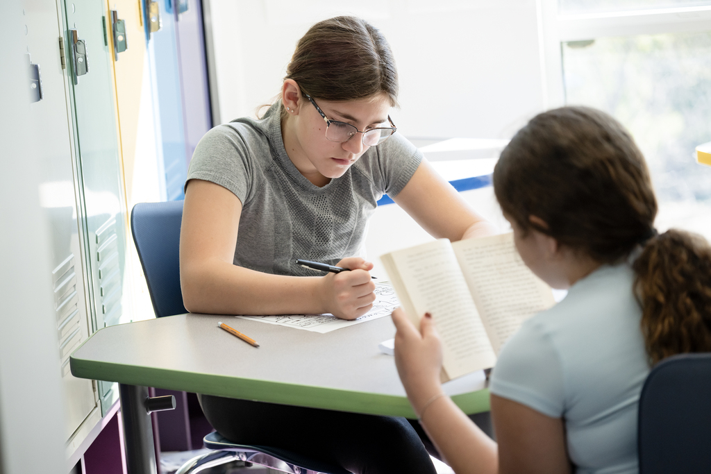 at a table, one student reads a book while another writes an assignment