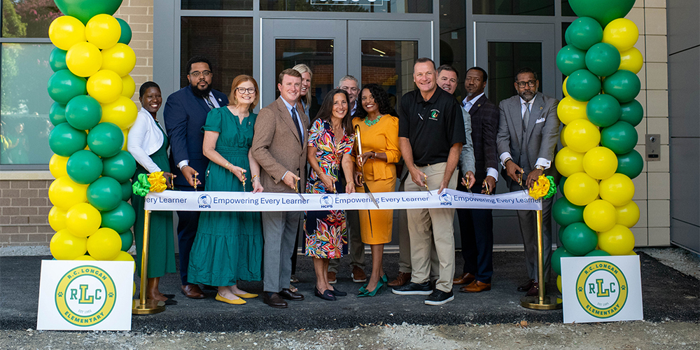 Leaders from HCPS and Henrico County pose at the ribbon-cutting ceremony for the new R.C. Longan Elementary School Building. They are holding scissors in front of a white HCPS ribbon that reads "Empowering Every Learner," and are surrounded by green and yellow balloons and signage for the school.