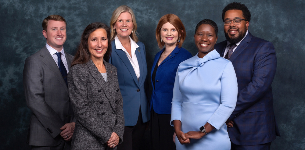 A group portrait of six school board members, four women and two men, standing together and smiling in formal business attire against a dark textured backdrop