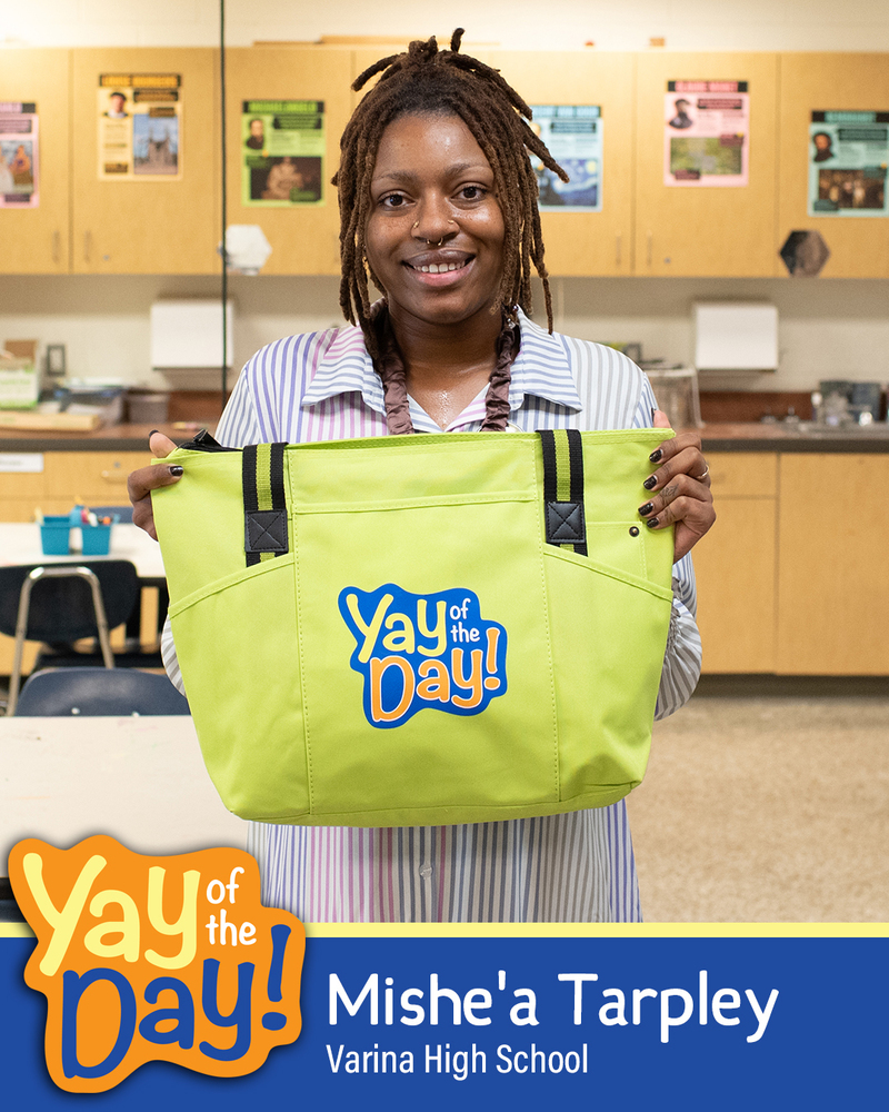 Mishe'a Tarpley with Varina High School smiles and holds up a green Yay of the Day tote bag in a classroom setting. Tarpley is identified as the Yay of the Day recipient.