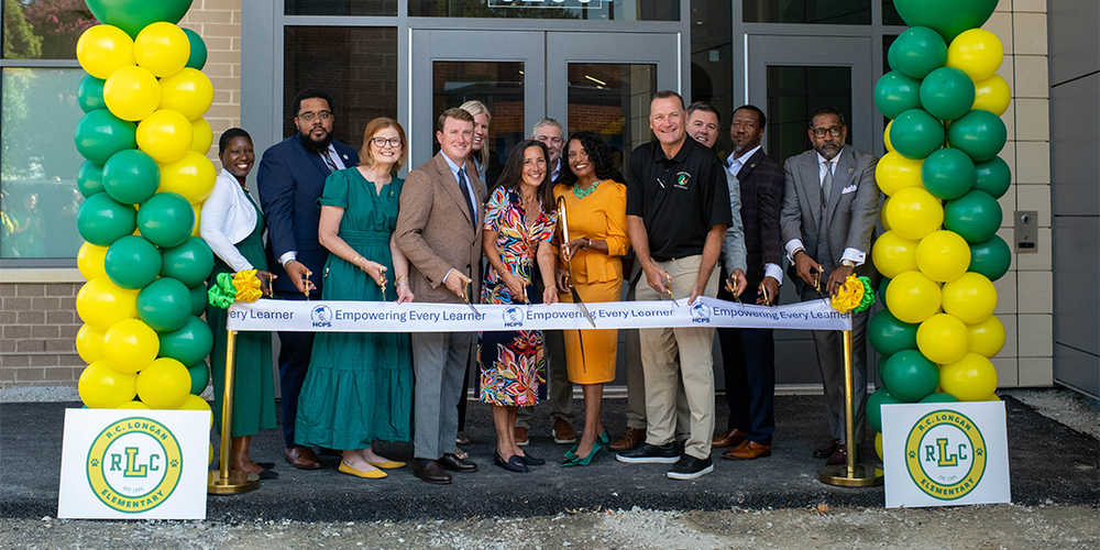 Leaders from HCPS and Henrico County pose at the ribbon-cutting ceremony for the new R.C. Longan Elementary School Building. They are holding scissors in front of a white HCPS ribbon that reads "Empowering Every Learner," and are surrounded by green and yellow balloons and signage for the school.
