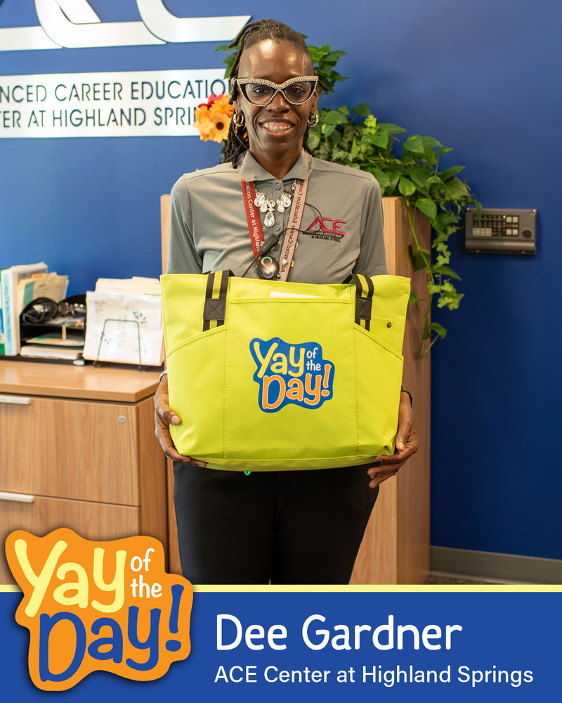 A woman smiles holding up a Yay of the Day bag in front of a school mural. The Yay of the Day recipient is identified as Dee Gardner of the ACE Center at Highland Springs.