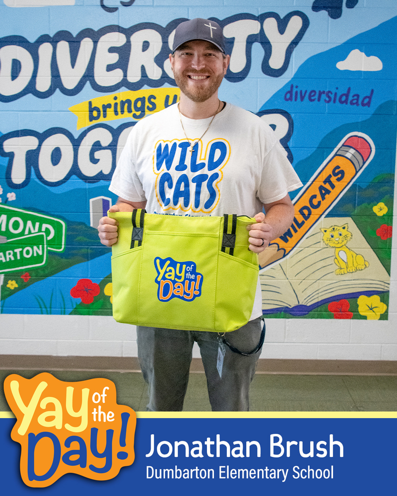 Jonathan Brush from Dumbarton Elementary School holding a yellow "Yay of the Day" tote bag in front of a colorful diversity-themed backdrop.