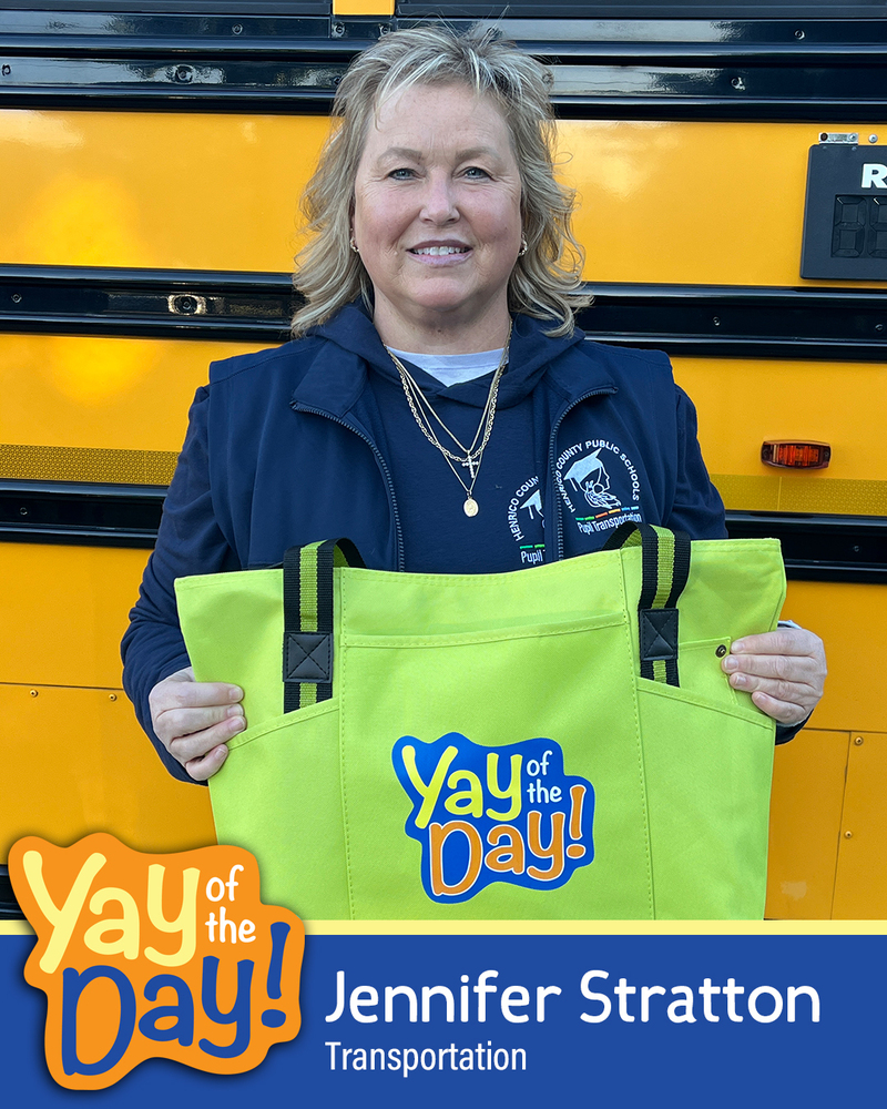 Bus driver Jennifer Stratton with Transportation holds up a Yay of the Day bag in front of a school bus for a Yay of the Day graphic.