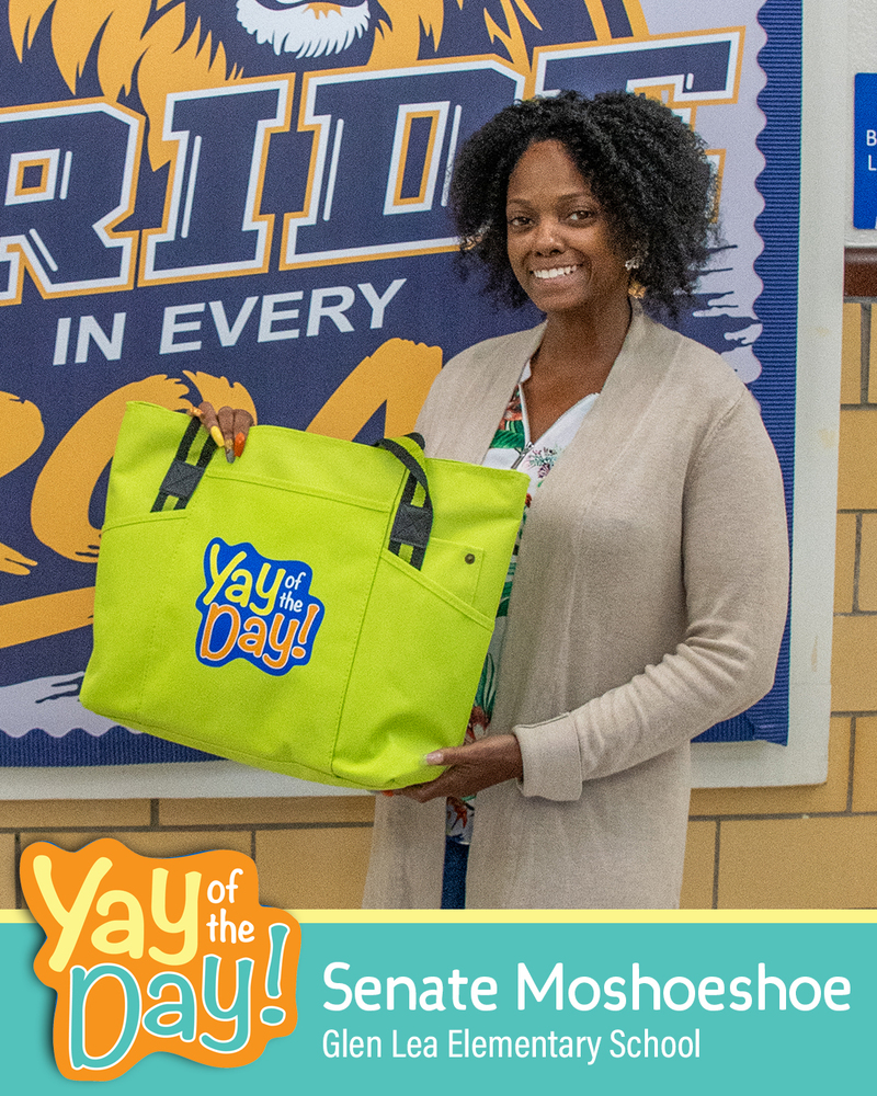 A woman smiles holding up a Yay of the Day bag in front of a school mural. The Yay of the Day recipient is identified as Senate Moshoeshoe, Glen Lea Elementary School.