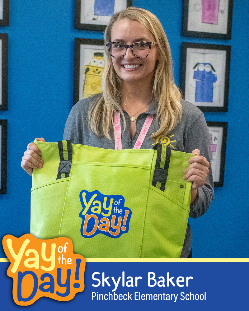 Skylar Baker from Pinchbeck Elementary School holding a yellow "Yay of the Day" tote bag in front of a blue wall with framed artwork.