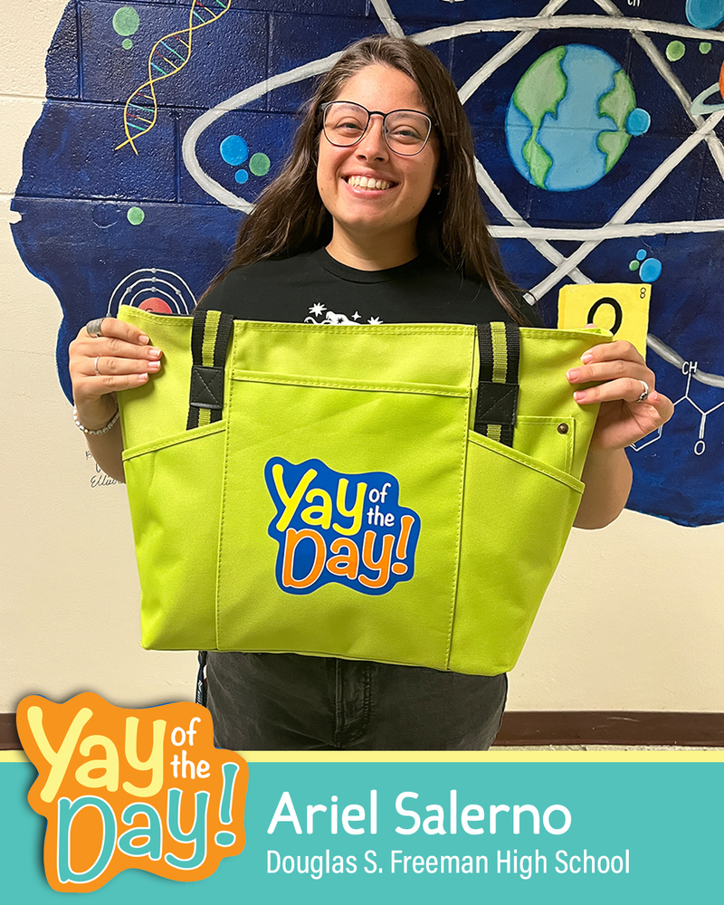 A woman with glasses smiles and holds up a Yay of the Day bag in front of a science-themed mural in a school hallway. The Yay of the Day recipient is identified as Ariel Salerno, Douglas S. Freeman High School.