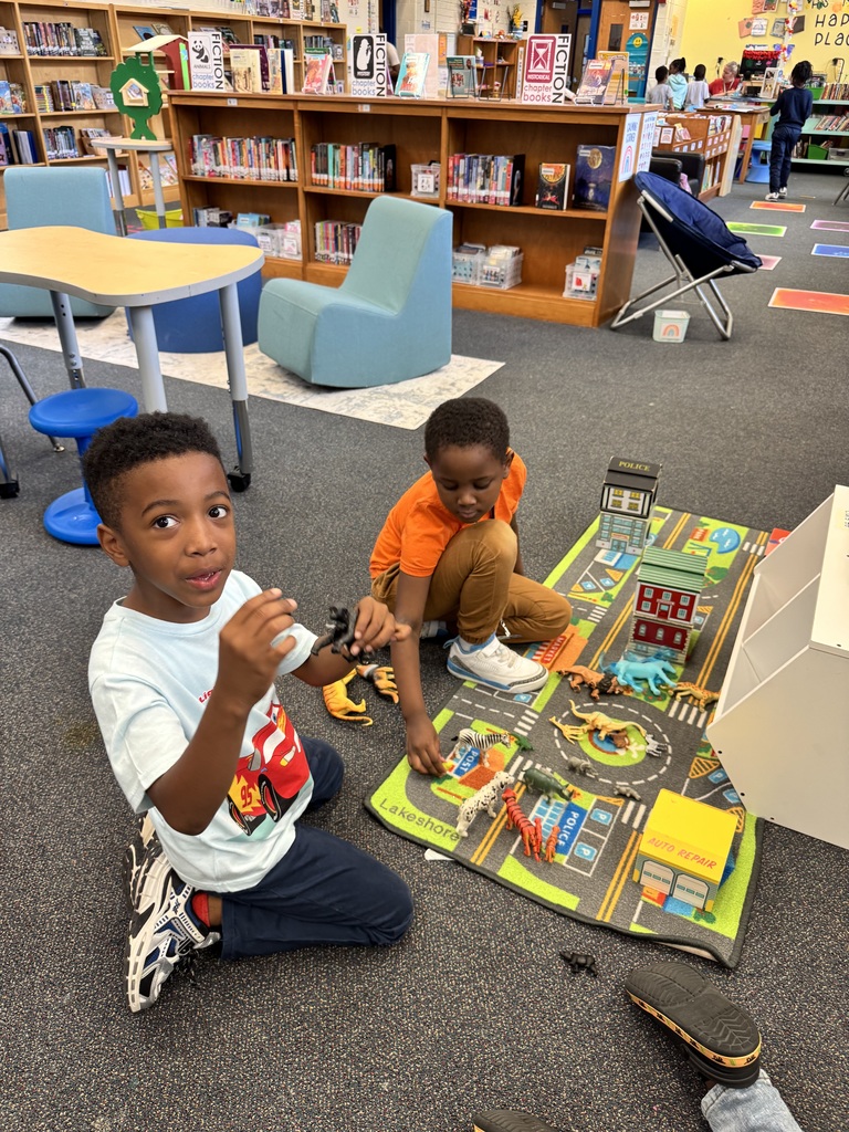 1st graders sit next to a car rug with animals and homes.