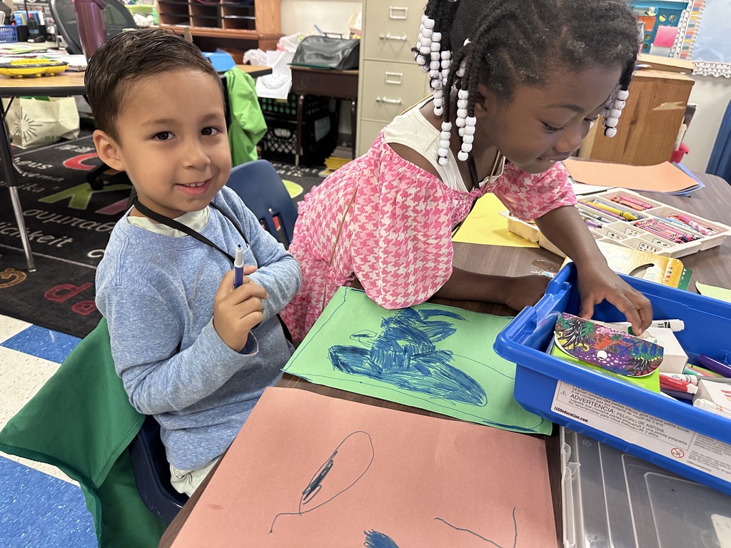 Two students color at their table. One is coloring with a blue marker while the other reaches for more supplies.