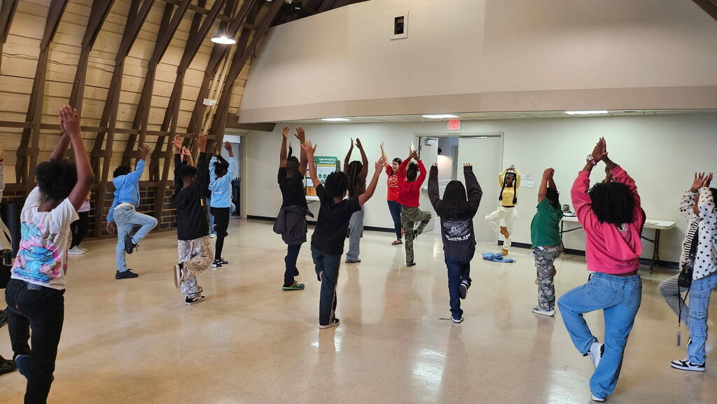 A class of students stands in a large room doing a yoga tree pose.