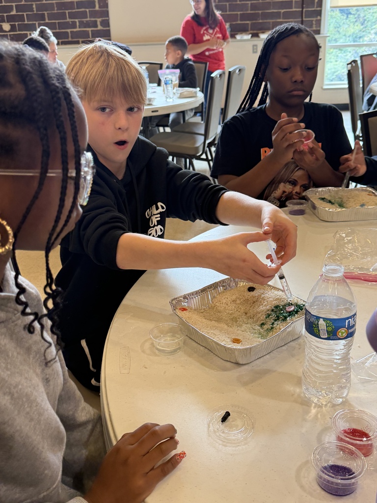 Three students sit at a table around a pan of rice while one uses a syringe to squirt liquid into the rice.