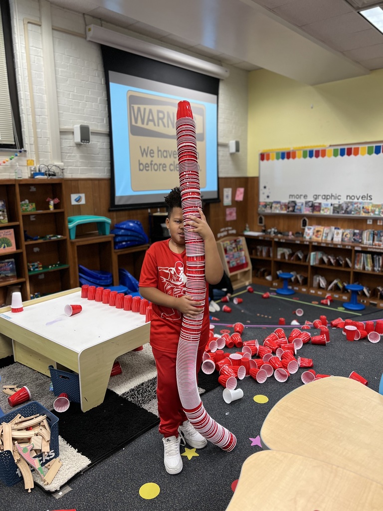 Student holds a very large stack of cups, taller than his head.