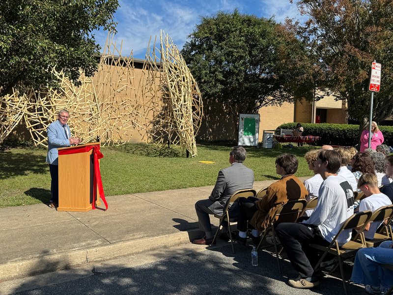 Henrico County Manager John Vithoulkas speaks in front of a wooden sculpture and a seated crowd outside of Godwin High School.