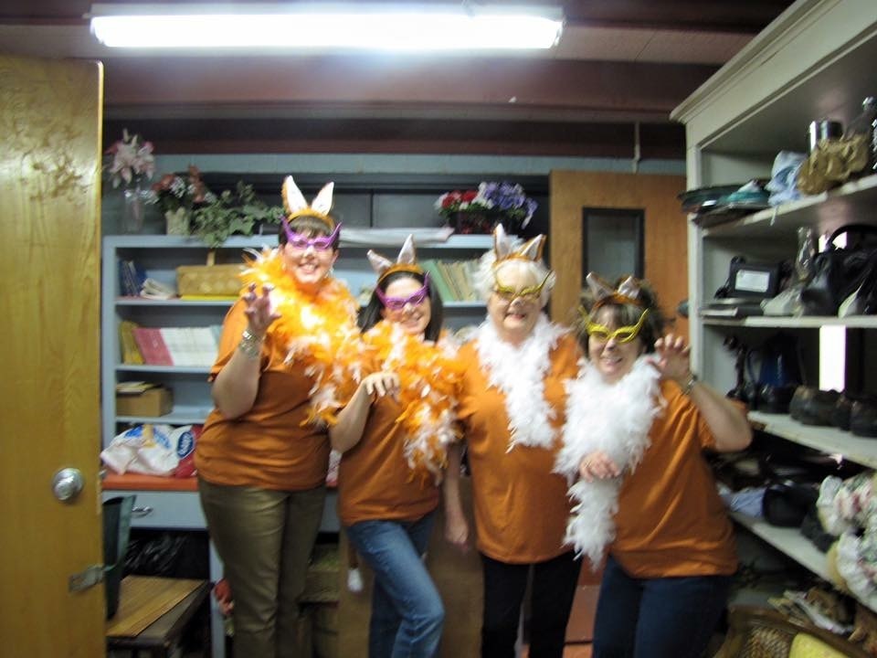 Four women wearing orange shirts and animal masks stand in a room with shelves and a door.