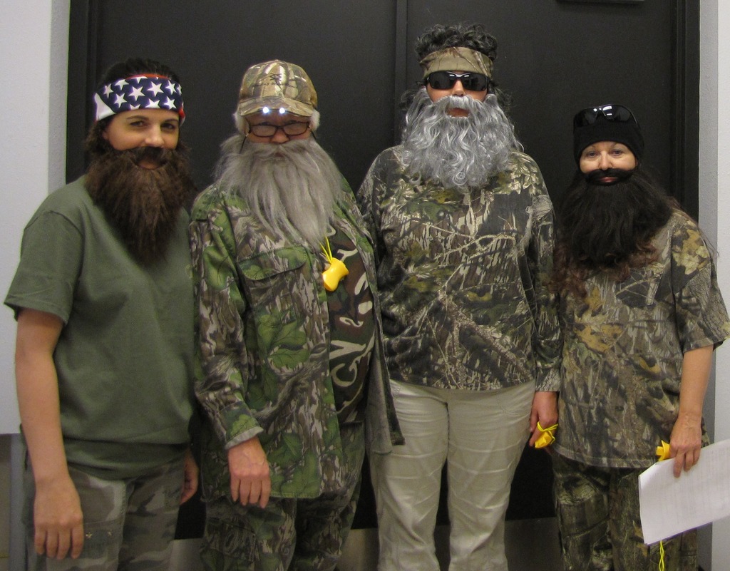 Four people dressed in camouflage clothing and beards pose against a black door. One holds a white paper.