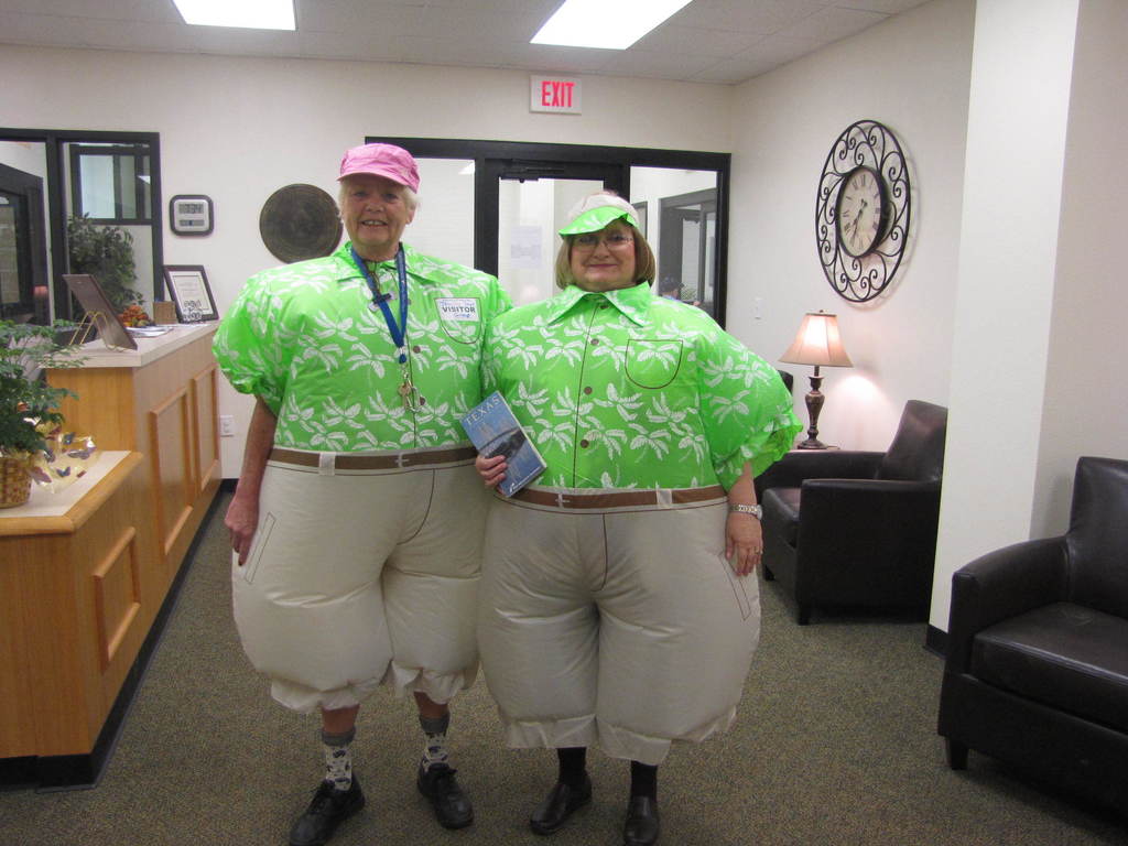 Two people in green shirts and white pants stand in an office with chairs, a desk, and a clock.