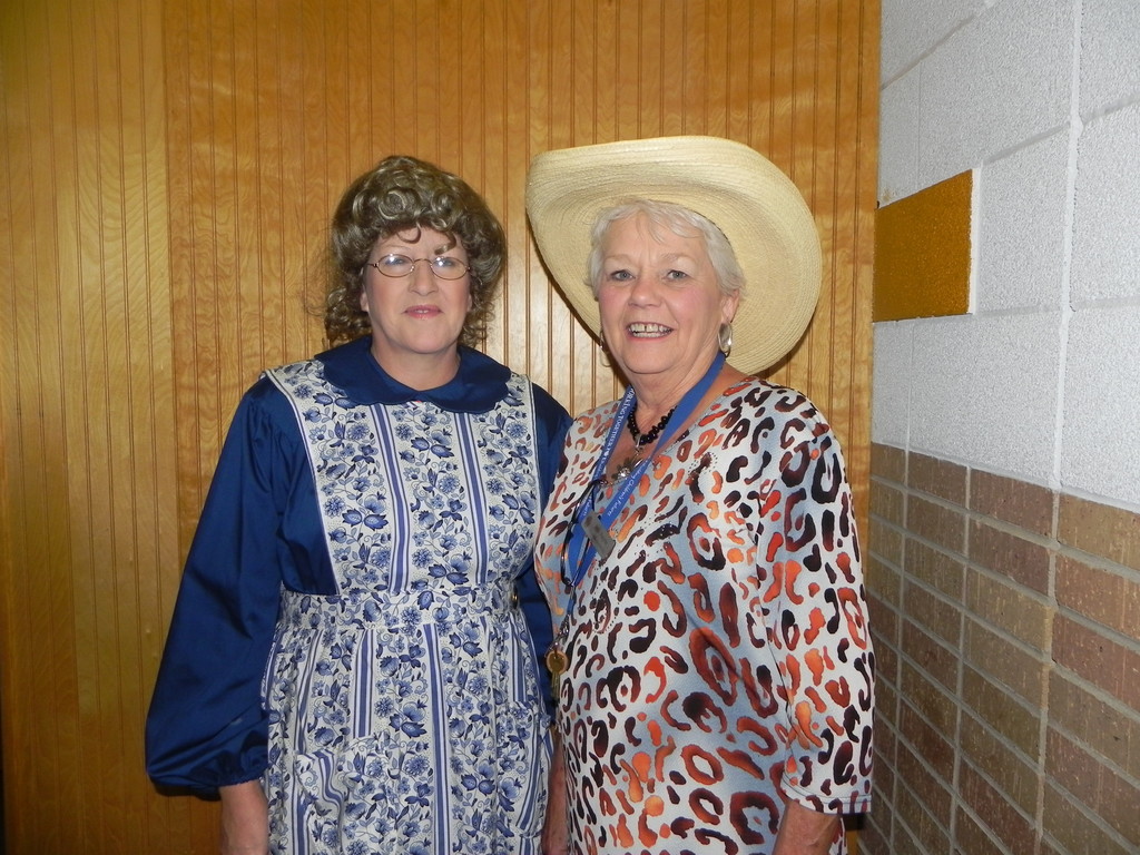 Two women standing next to each other. One wears a blue dress and a cowboy hat, the other wears glasses.