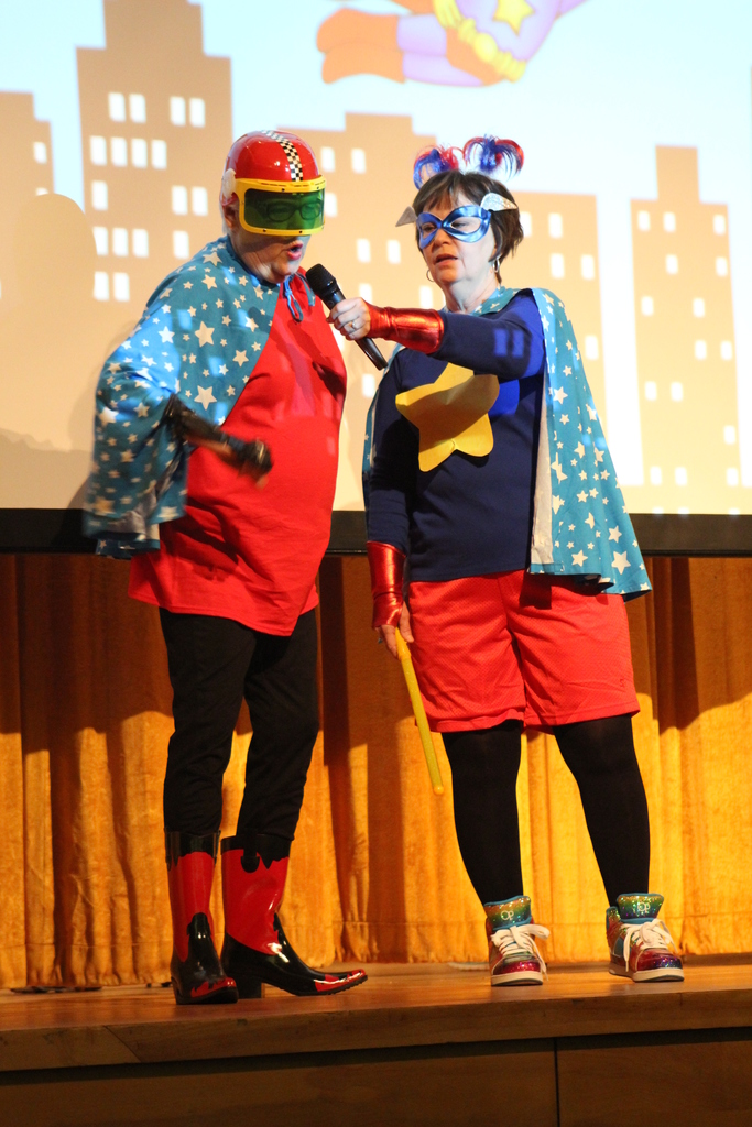 Two individuals in superhero costumes stand on a stage, holding props and microphones, with a projector screen in the background.