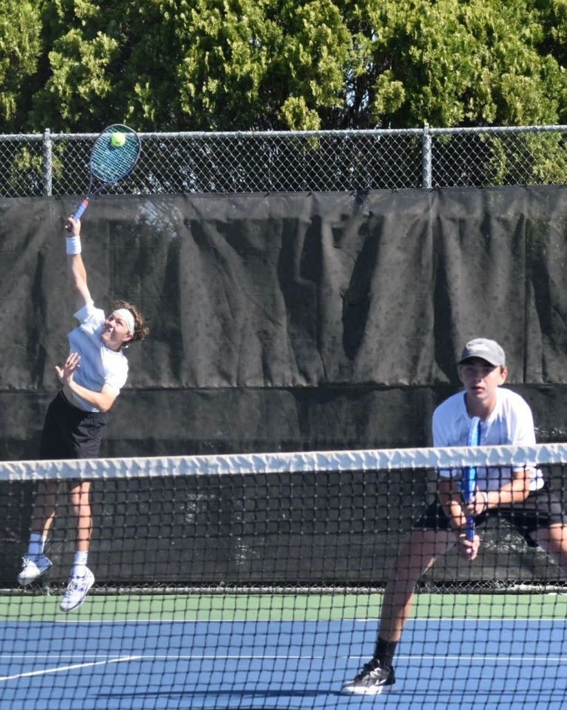 Two tennis players on a court, one leaping to hit a ball, another holding a racket, with a net and trees in the background.