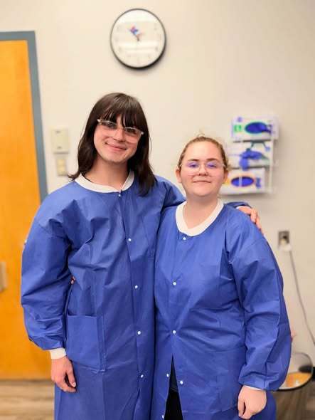 Two individuals wearing blue lab coats stand close together in a room with a yellow door.