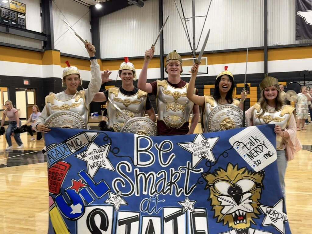 A group of people dressed in costumes pose for a picture with a banner in a gym.