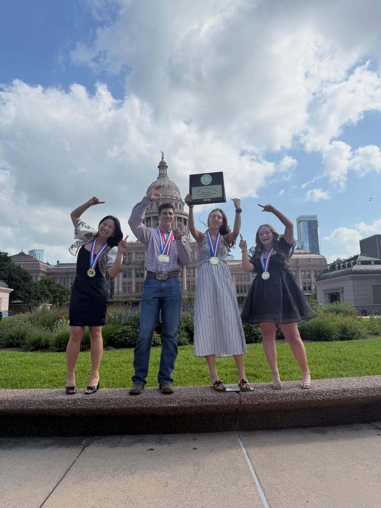 Four people pose for a photo with awards on a concrete walkway, holding a framed certificate, with a grassy area and city skyline in the background.