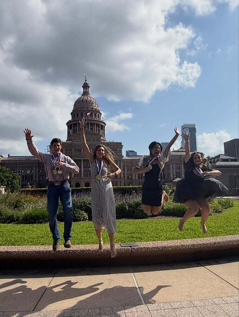 Four people jump and celebrate in front of a grassy area with a concrete barrier.